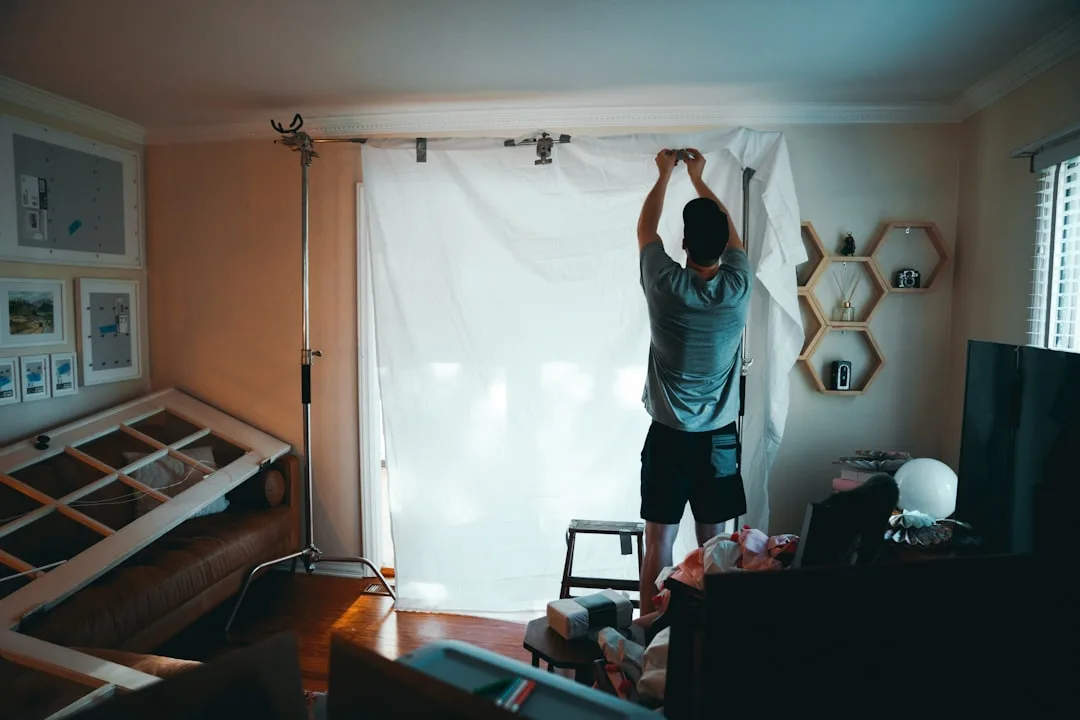 Man hanging wide white fabric panel on curtain rod using light stand support for full luxurious drapery look