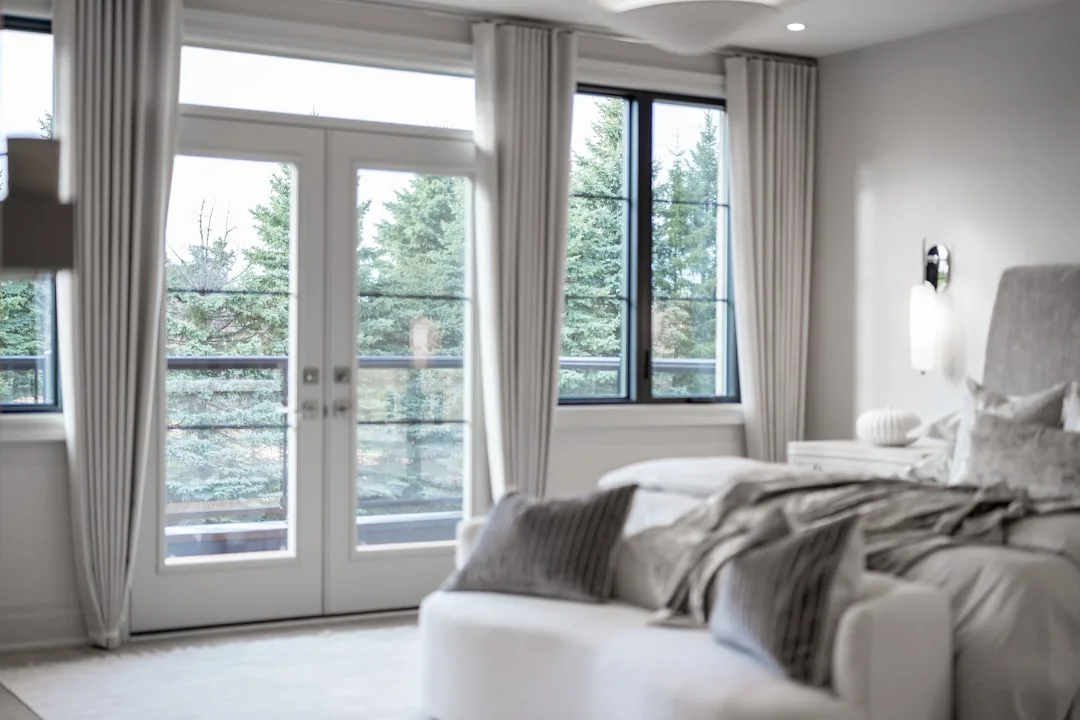 Layered bedroom curtains with sheer drapes over French doors allowing natural light in modern white bedroom