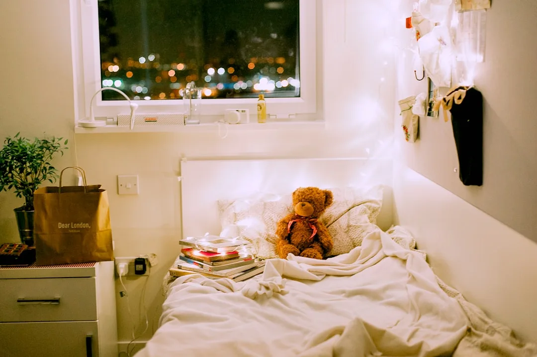 Cozy dorm room at night with fairy lights, teddy bear, stacked books on bed, and city lights visible through window