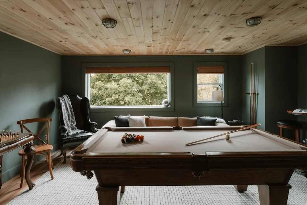 Cozy basement game room with pool table, wood-plank ceiling, dark green walls, and natural light from large windows