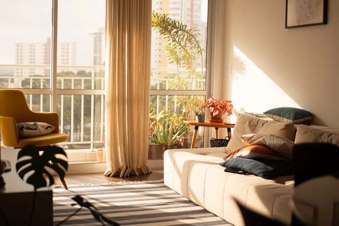Sunlit apartment living room with white sofa, layered cushions, yellow chair, sheer curtains and balcony plants