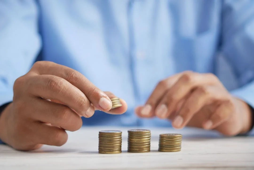 Hands stacking gold coins into three growing piles on white surface, symbolizing budget allocation and savings strategy