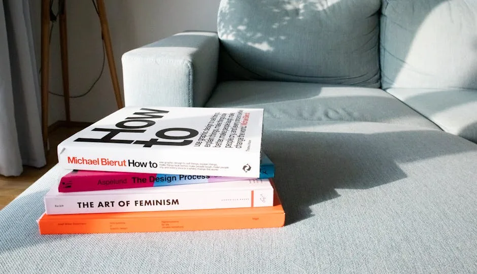 Stack of design and lifestyle books on a light grey sofa in a minimalist living room with natural sunlight