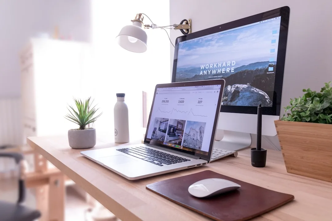 Organized home office desk with MacBook, iMac monitor, plant, water bottle, and Apple Magic Mouse on wooden surface
