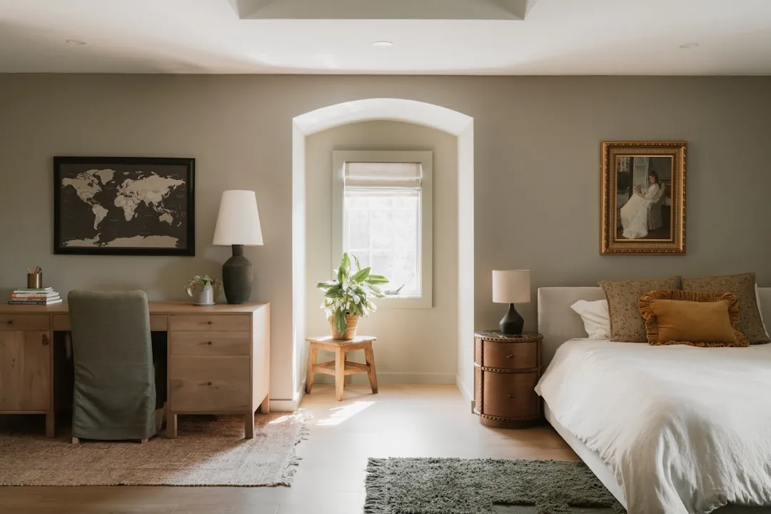 Bedroom with arched window nook styled with potted plant and wooden stool, flanked by desk and bed in neutral tones