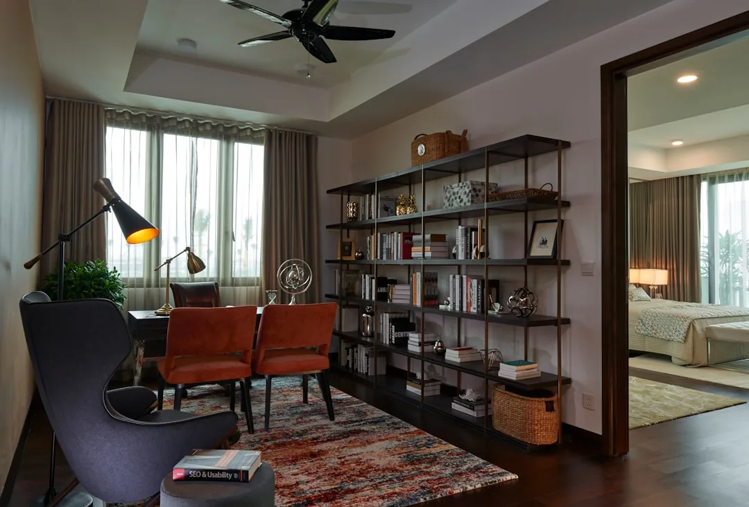 Home office zone with bookshelf divider, desk, orange chairs, and adjacent bedroom visible through open doorway