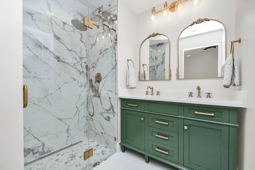 Luxury bathroom remodel with green vanity, gold fixtures, marble shower tiles, and arched mirrors under $10000 budget