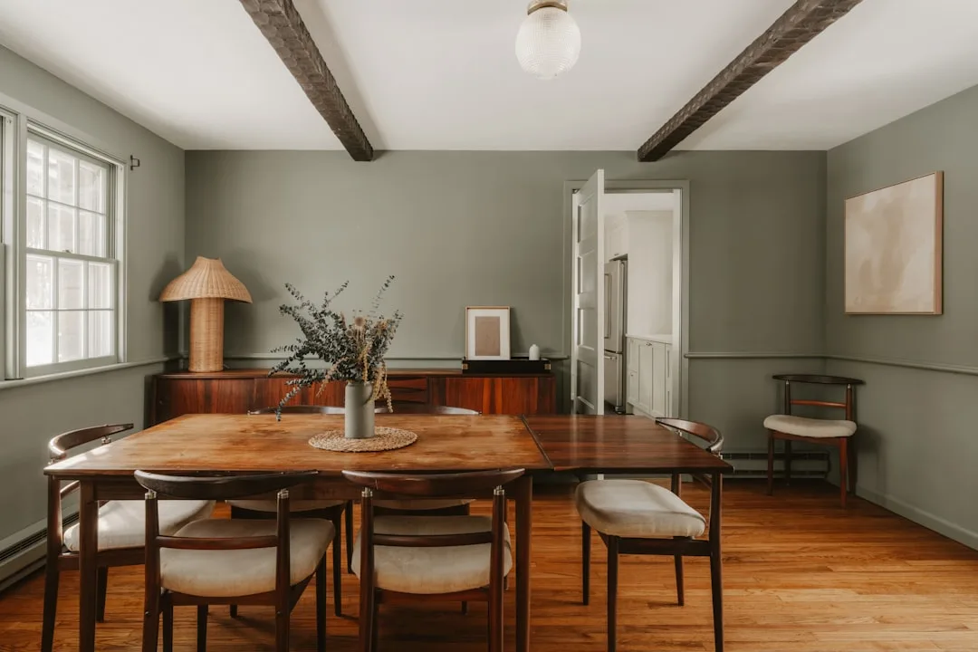 Mid-century modern dining room with warm brown wood table and chairs against sage green walls with exposed ceiling beams