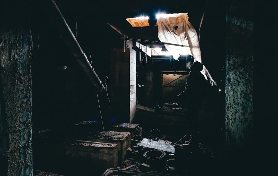 Dark unfinished basement with debris, exposed pipes, and a silhouetted person exploring the space with a light