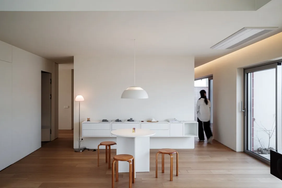 Minimalist Japandi dining area with white pedestal table, wooden stools, pendant lamp, and clean white cabinetry on ligh