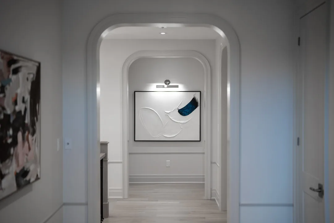 Hallway gallery wall lit by a dedicated picture light above framed abstract art in a white corridor with arched doorway