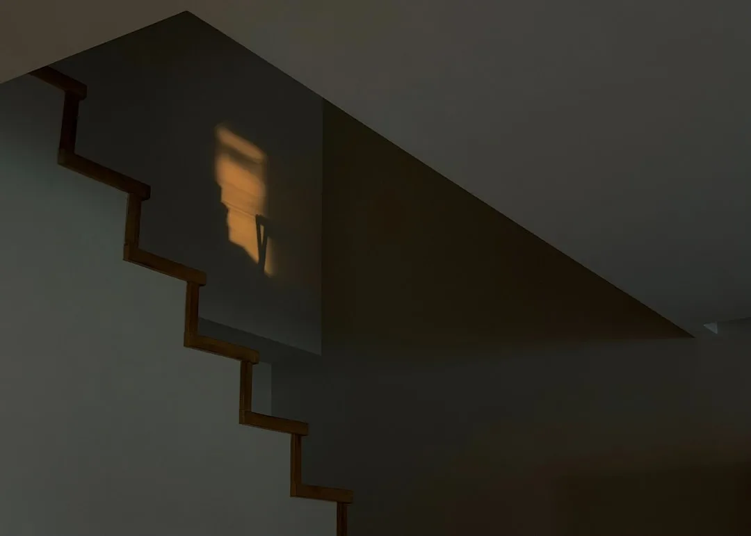 Warm lamp light projection casting shadow on staircase wall creating natural lighting zones in dim interior space