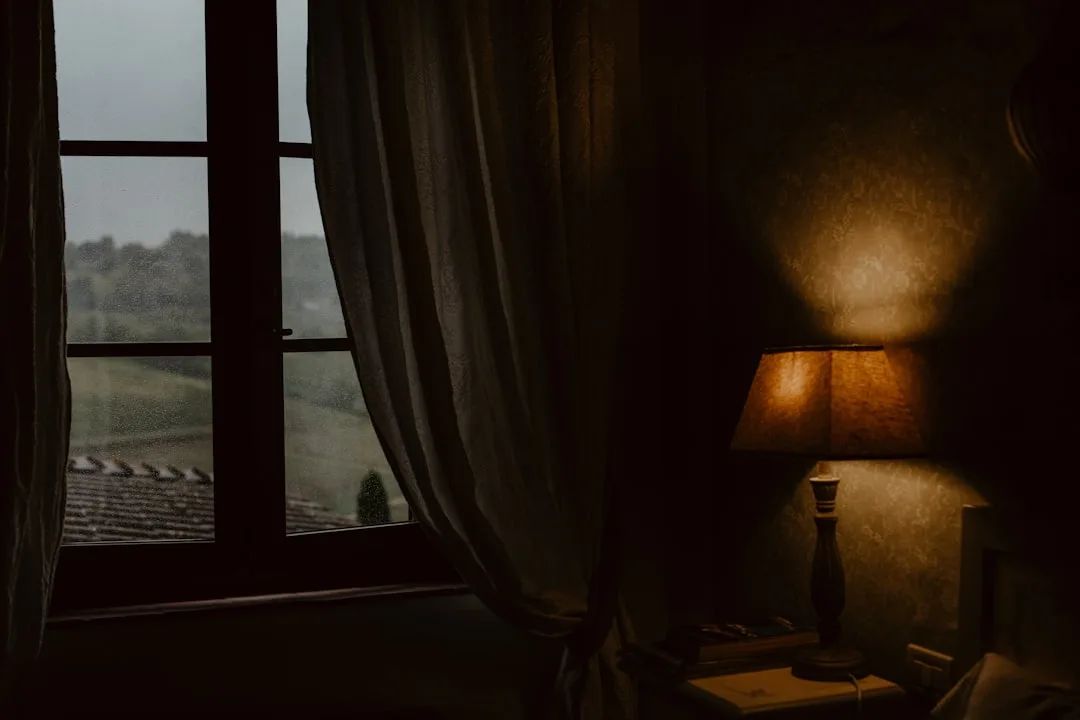 Warm amber bedside table lamp glowing in dark bedroom corner beside rainy window with curtains