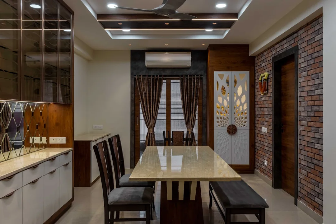 Modern dining room with marble table, dark wood chairs, decorative wall panel, brick accent wall and false ceiling light