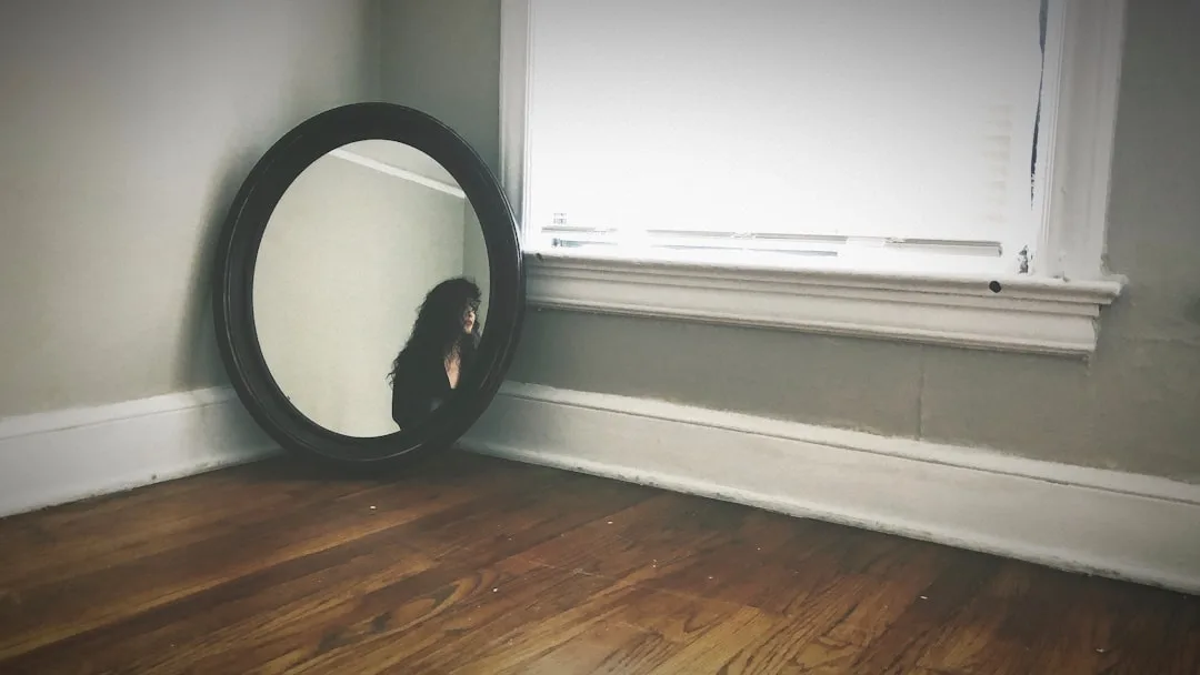Oval black-framed mirror leaning against wall in empty room reflecting person, next to bright window on hardwood floor