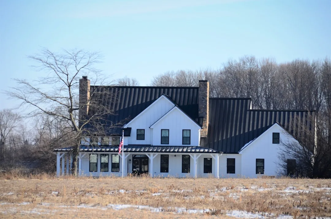 Modern farmhouse with white board-and-batten siding, black metal roof, stone chimneys, and wraparound porch in rural win
