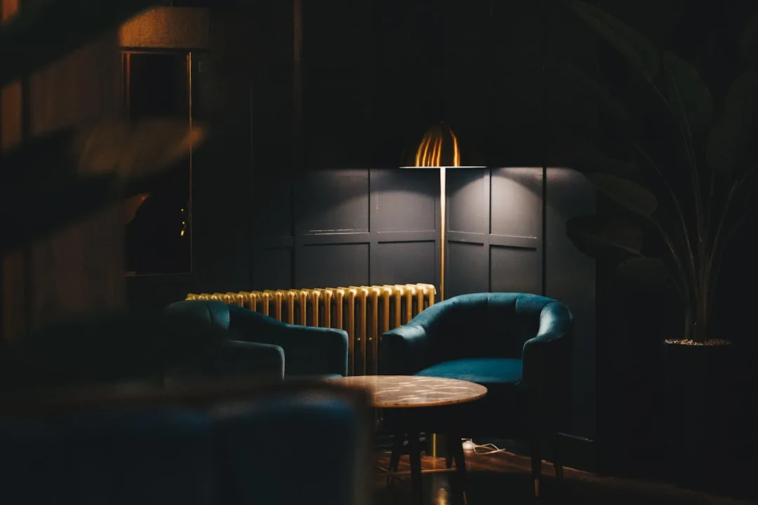 Moody lounge with teal velvet armchairs, gold floor lamp casting warm focused light against dark paneled wall