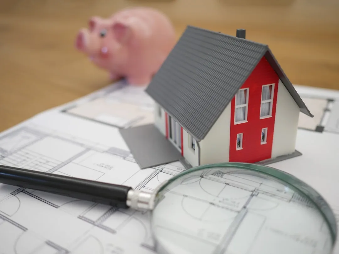 Miniature house model on architectural blueprints with magnifying glass and piggy bank symbolizing home permit inspectio