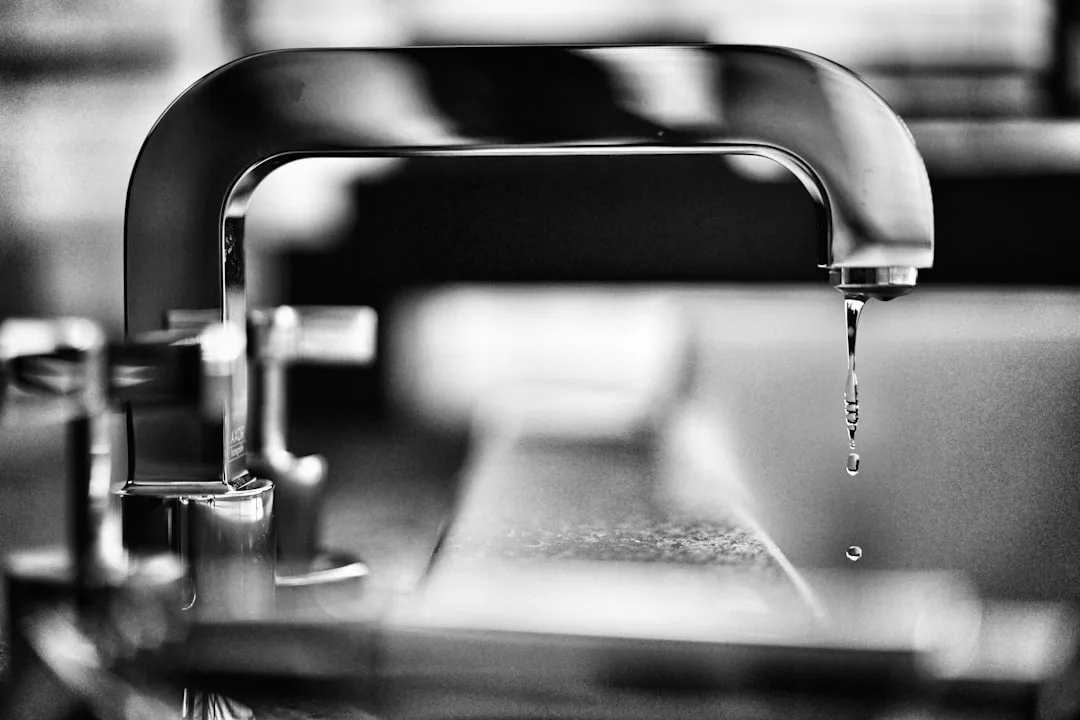 Close-up black and white photo of a modern chrome kitchen faucet dripping water onto a sink surface