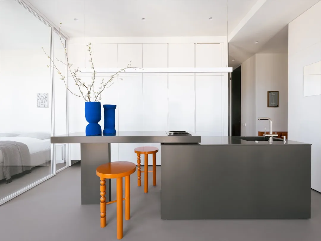 Minimalist studio apartment with gray kitchen island, orange stools, blue vases, and open bedroom area beyond glass part