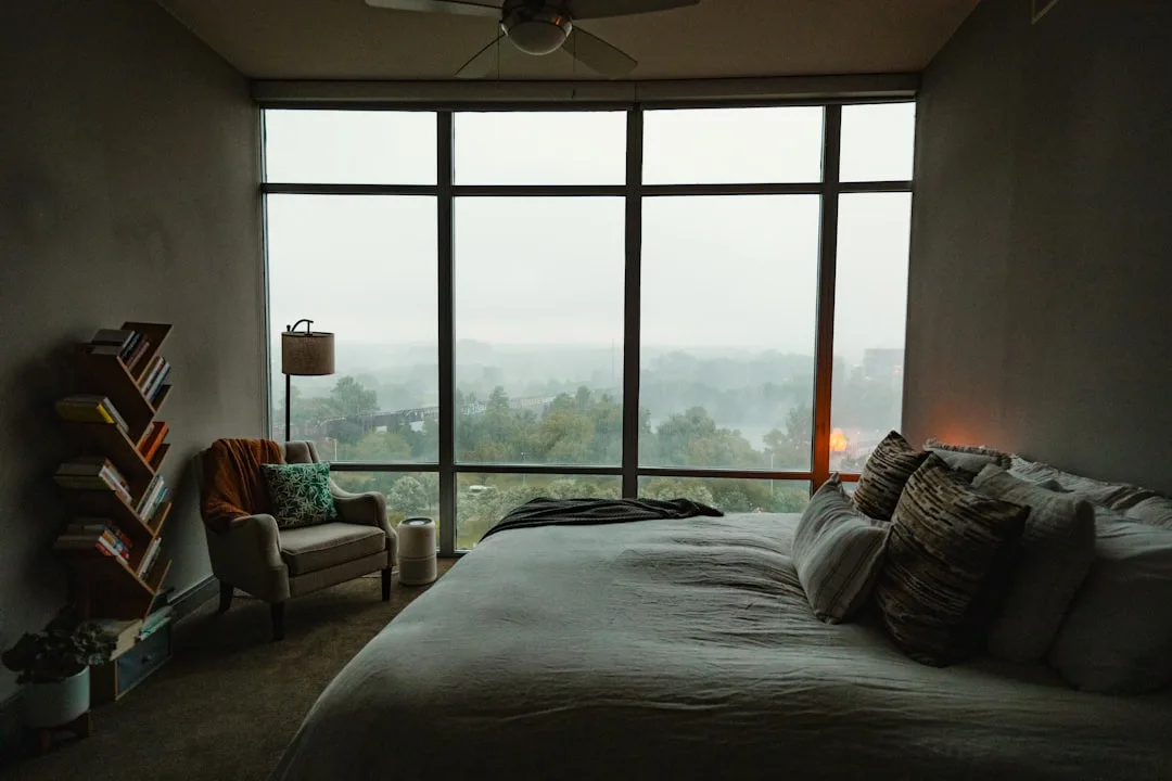 Moody bedroom with floor-to-ceiling windows, neutral linen bedding, wood bookshelf, and misty forest view
