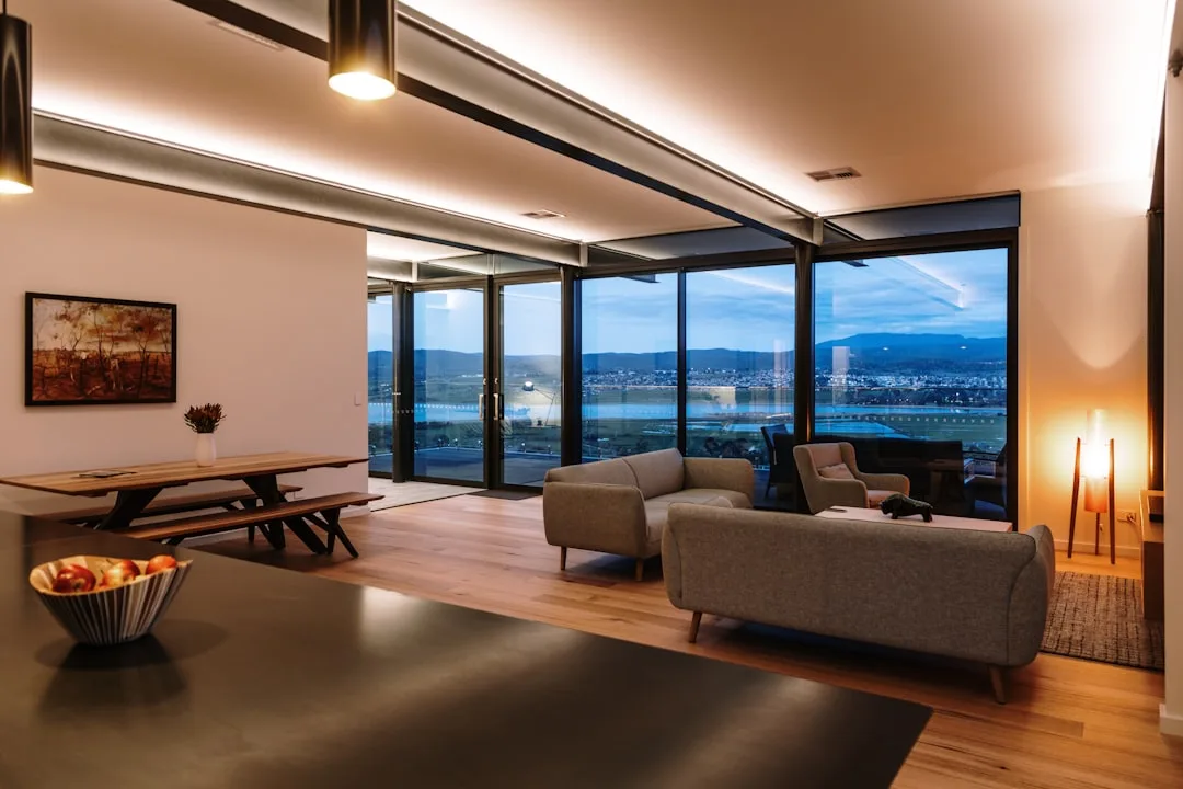 Modern living room with warm accent lighting, cove ceiling lights, floor lamps, and floor-to-ceiling windows at dusk