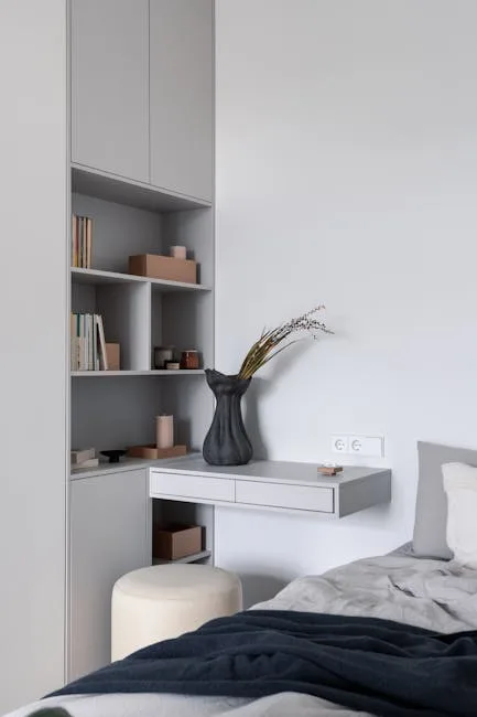 Small bedroom nook with floating wall desk, built-in grey shelving, black vase, and stool in minimalist bedroom