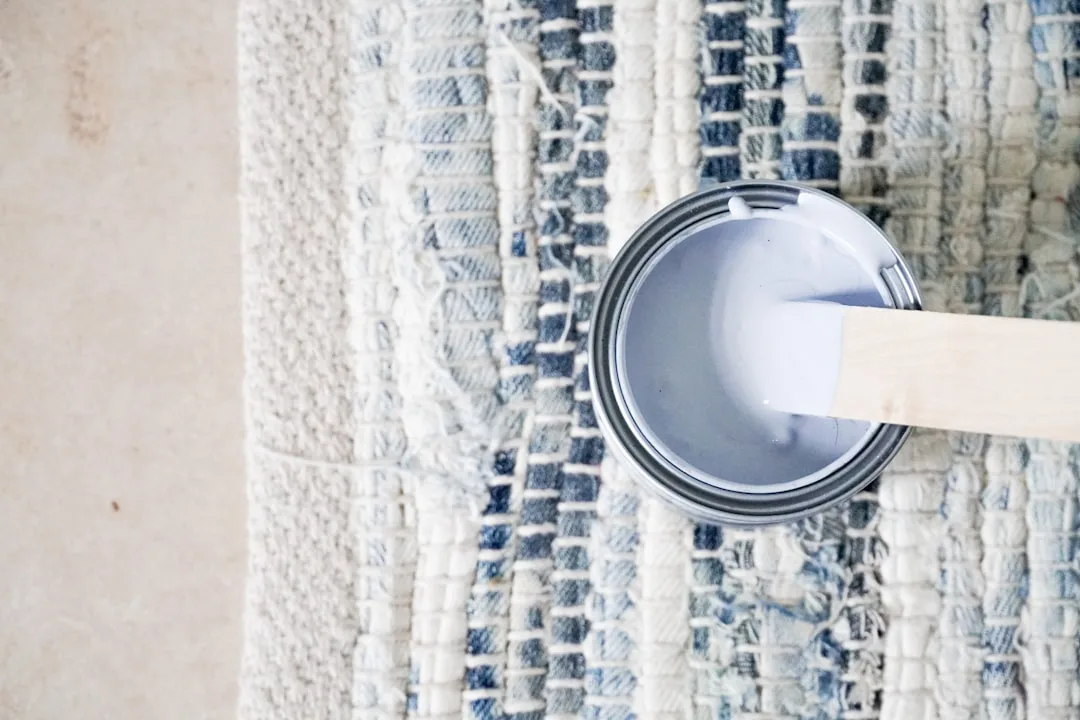 Open paint can with white paint and wooden stirrer resting on a blue and white woven rug
