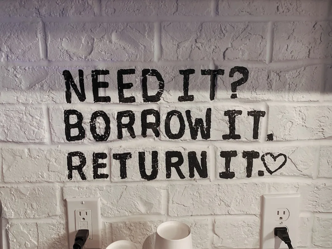 Bold black stenciled text on white painted brick wall reading Need It Borrow It Return It with heart symbol