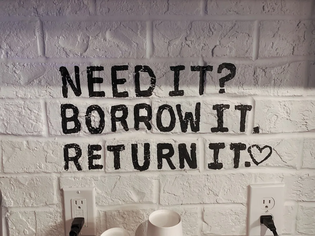 Black stenciled text on white painted brick wall reading Need It? Borrow It. Return It. with a heart symbol