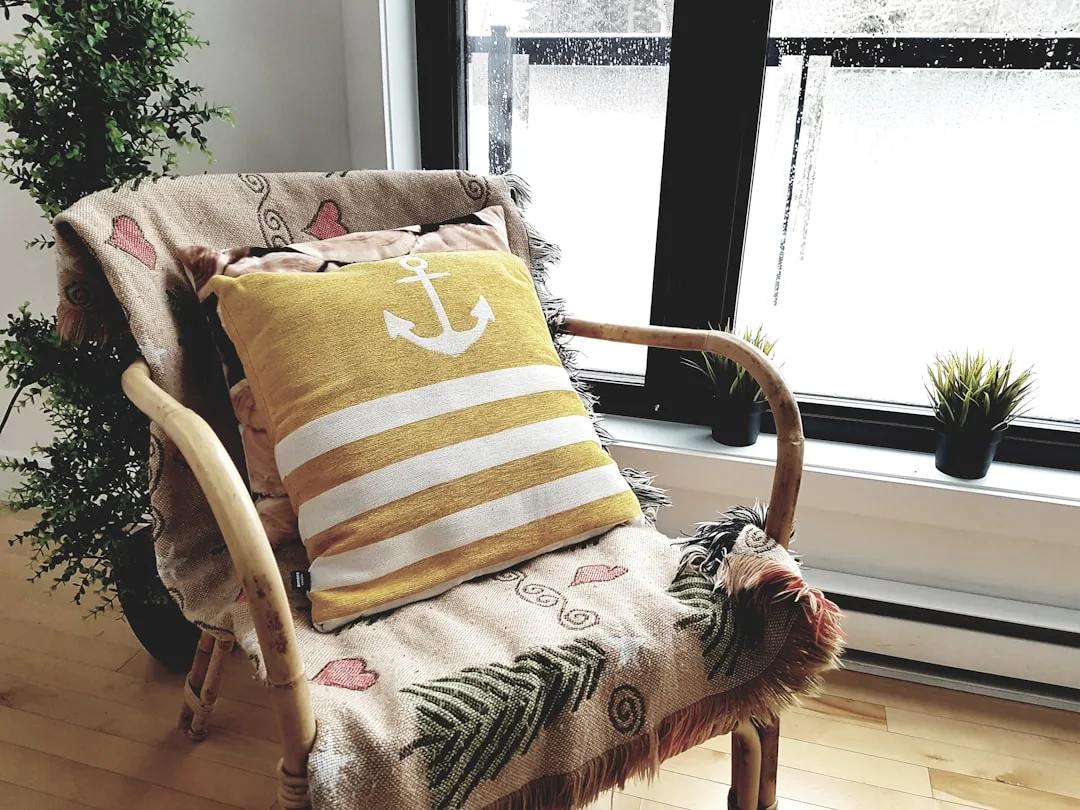 Yellow nautical anchor pillow on rattan chair with patterned throw blanket near window in small apartment