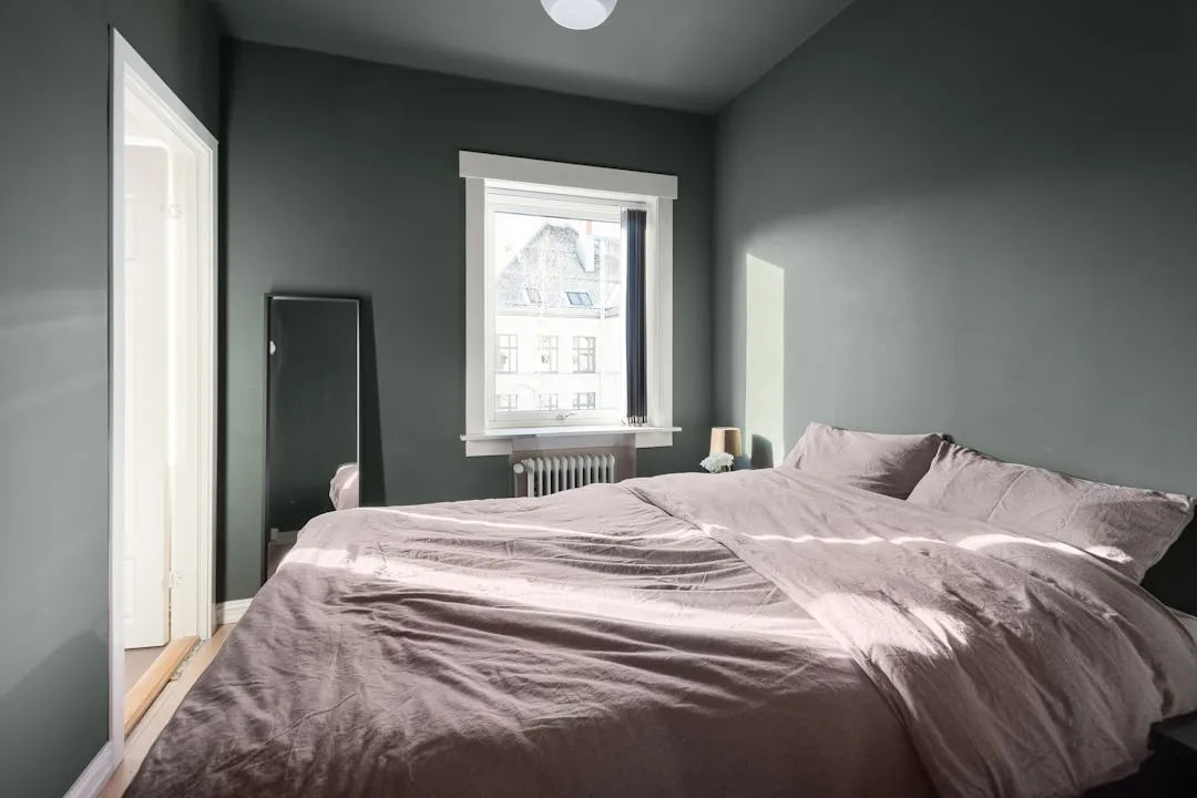 Bedroom with dark sage green walls, unmade linen bed, floor mirror, and natural window light from urban view