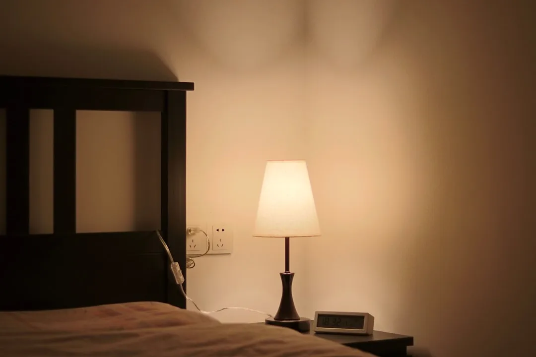Warm glowing bedside table lamp on nightstand next to alarm clock in dark bedroom ambient lighting
