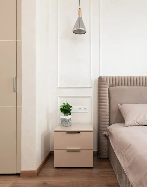 Beige two-drawer nightstand alternative beside upholstered bed in small bedroom with pendant light and small plant