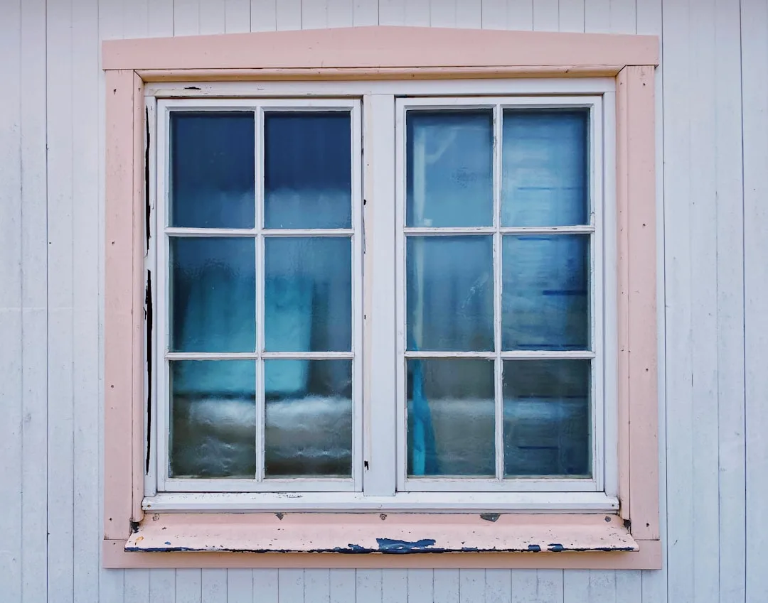 Peeling paint on pink wooden window frame with frosted blue glass panes showing common window maintenance mistakes