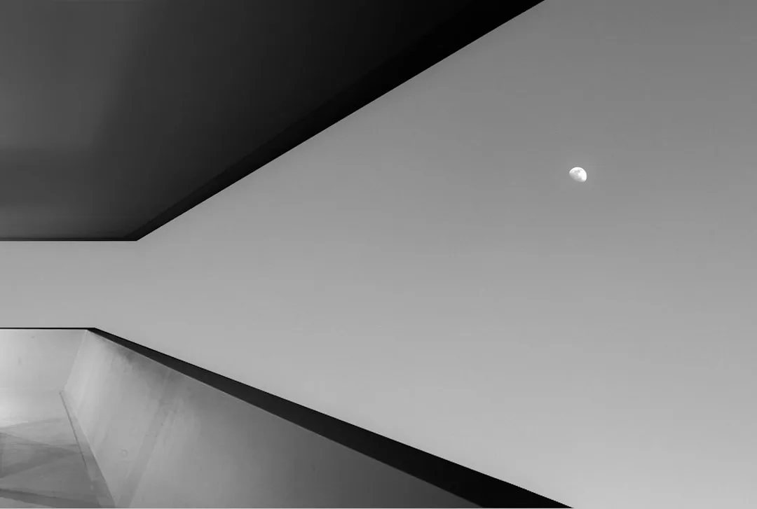 Black and white architectural photo showing dramatic ceiling height and geometric lines with moon visible through openin
