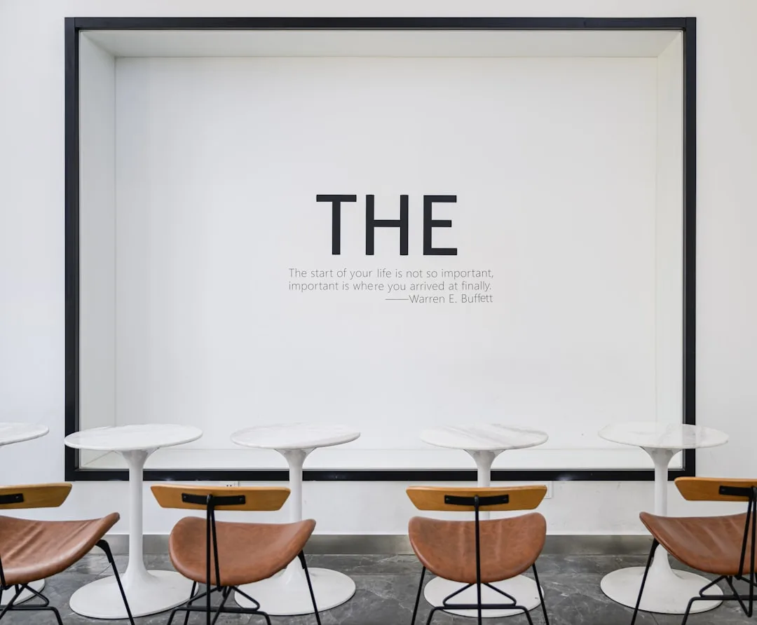 Minimalist cafe interior with white tulip tables, brown leather chairs, and Warren Buffett quote on white wall