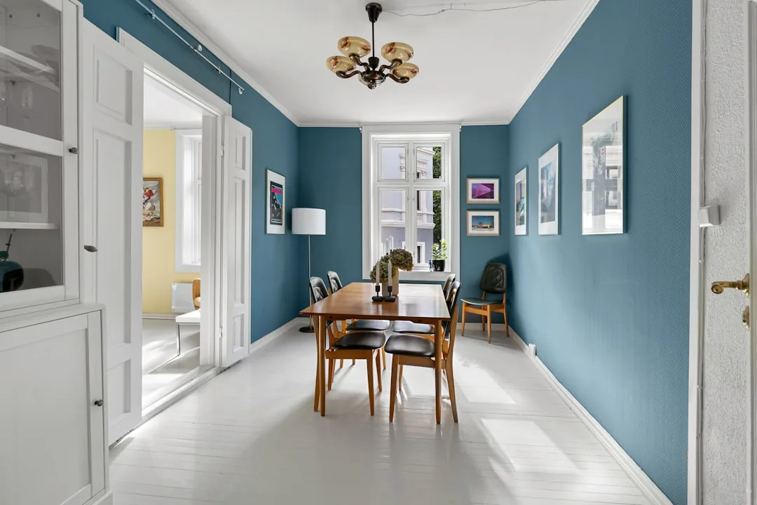 Long narrow dining room with blue walls, wooden mid-century table and chairs, white painted floors and chandelier