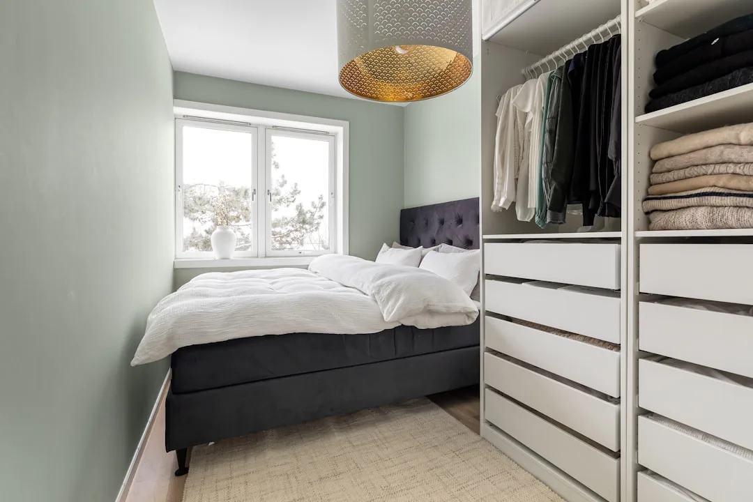 Small bedroom with open wardrobe system showing hanging clothes and folded items beside a dark upholstered bed