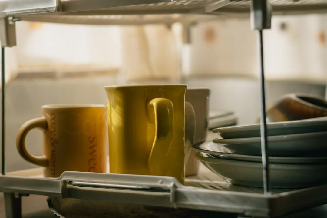 Yellow and olive-toned ceramic mugs on a kitchen shelf alongside stacked gray plates and bowls
