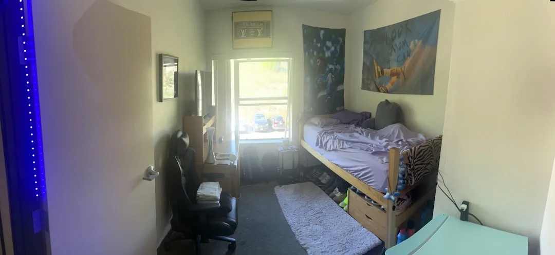 College dorm room with lofted bed, gaming chair, desk setup, blue LED lights, and sports poster wall decor