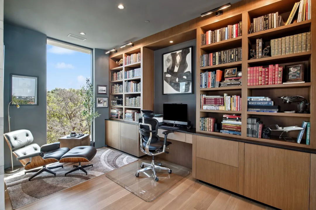 Modern home office with built-in wood bookshelves, Eames lounge chair, ergonomic desk chair, and floor-to-ceiling window