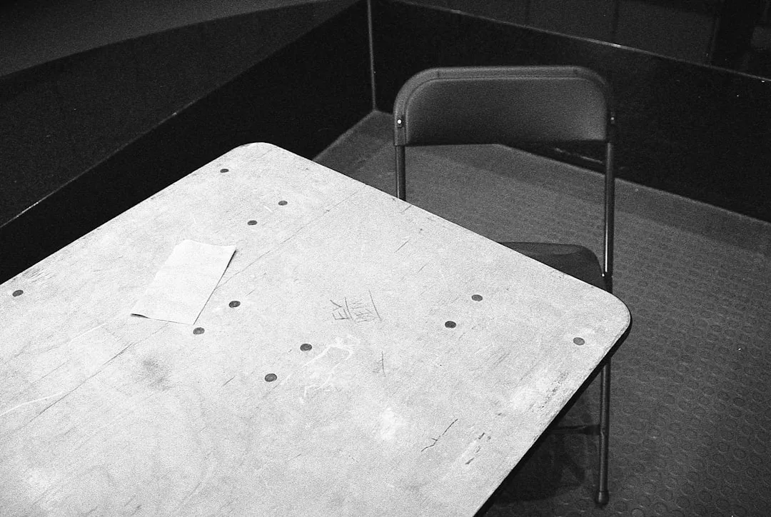 Worn institutional folding table and metal chair in a dorm room corner, black and white photo