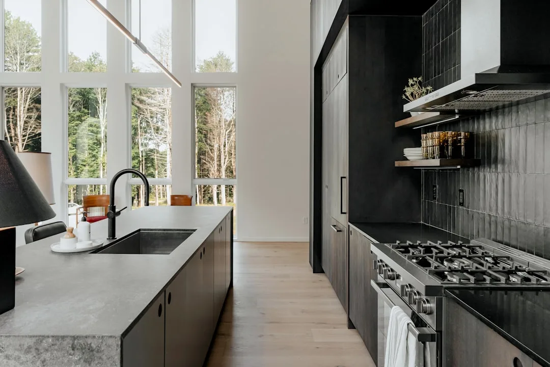 Modern dark kitchen with professional gas range, black tile backsplash, stone island countertop, and floor-to-ceiling wi