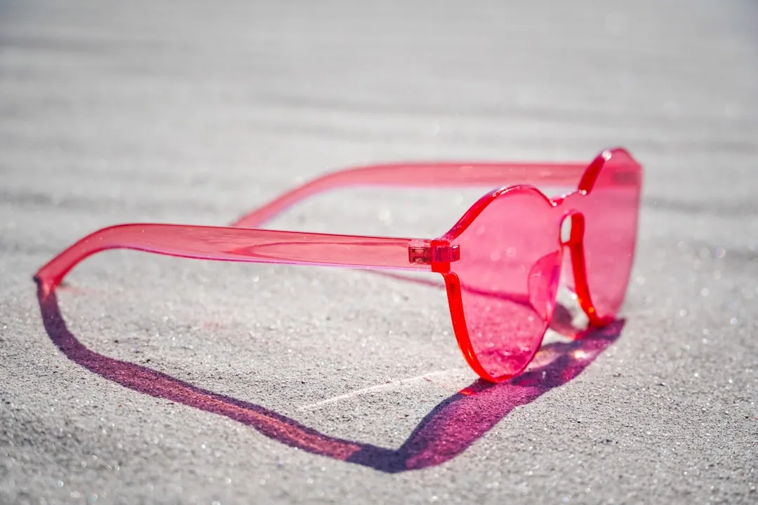 Translucent pink red sunglasses resting on sandy beach casting colorful shadow in bright sunlight