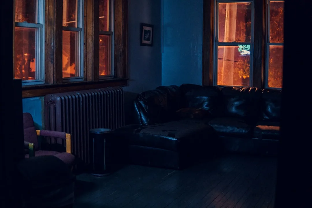 Dark moody living room at night with leather sofa, cast iron radiator, and warm orange glow from windows