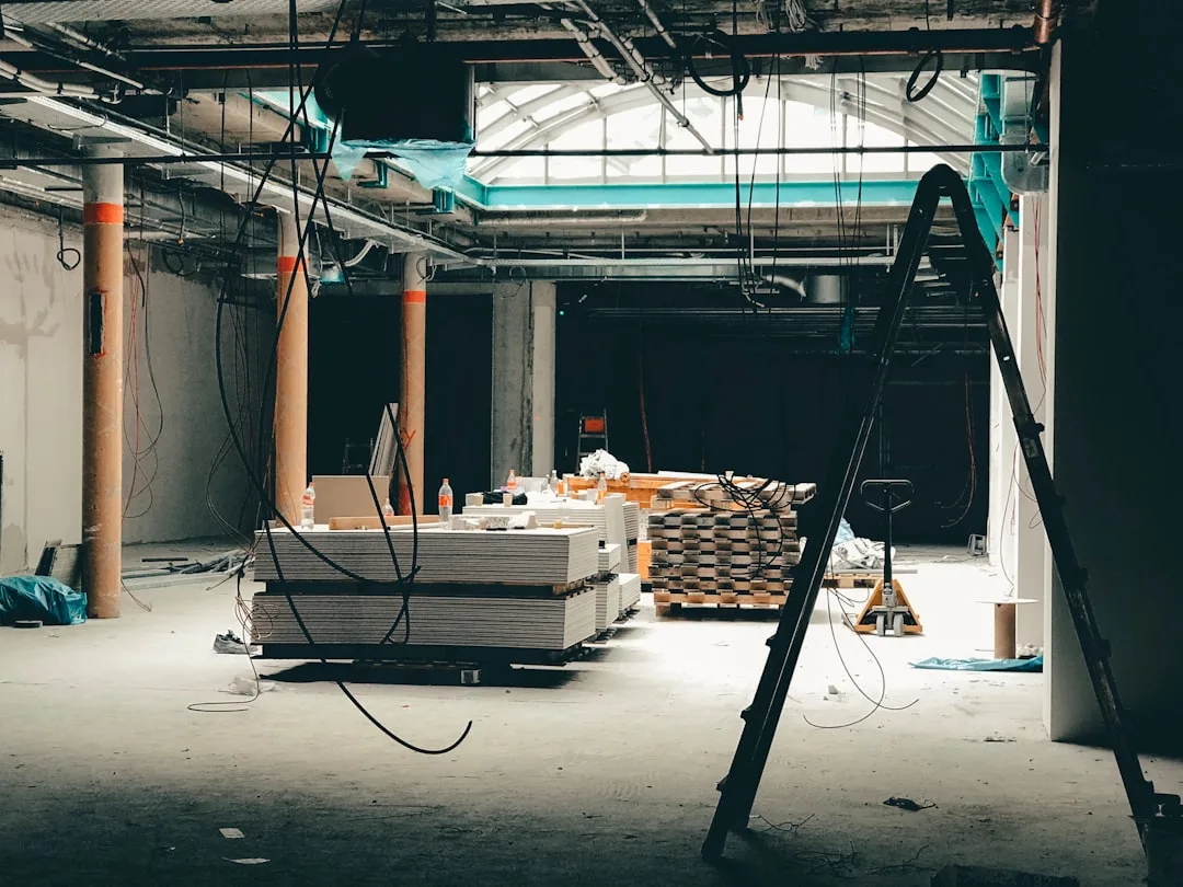 Interior construction site with stacked drywall, hanging electrical wires, ladders and building materials on concrete fl