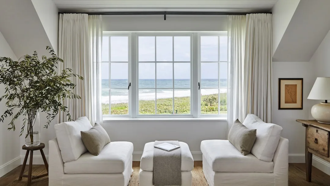 White linen curtains hung high near ceiling on black rod above large triple window with ocean view in coastal living roo