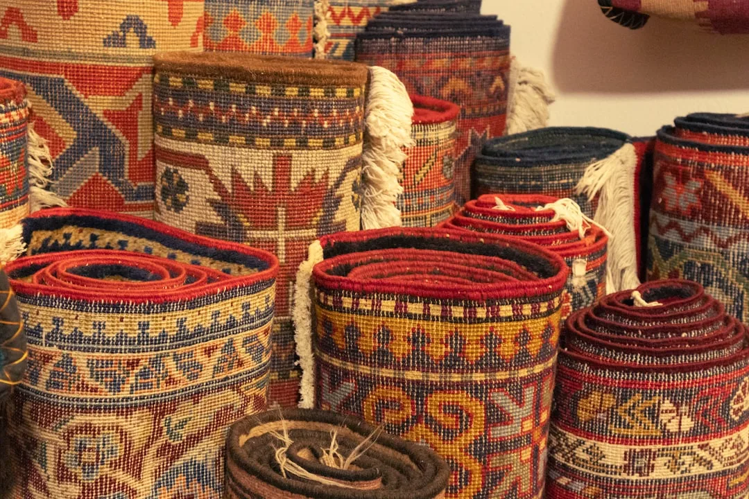 Multiple rolled traditional Persian and kilim rugs stacked together showing colorful geometric patterns and various size