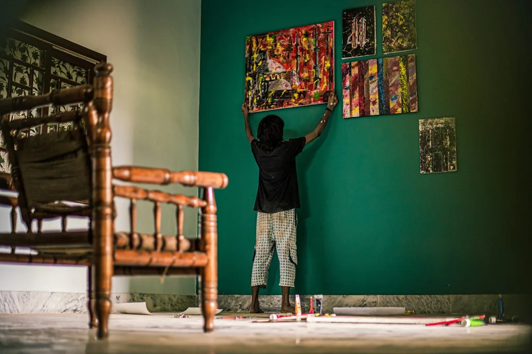 Person hanging colorful abstract paintings on a teal focal wall in a room, demonstrating DIY wall art arrangement techni